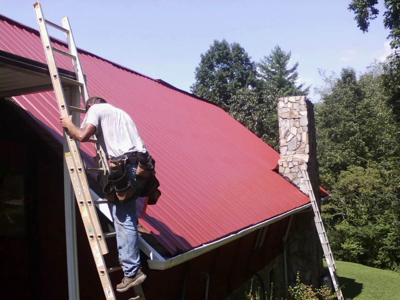 Roofer on a ladder beside a red metal roof and stone chimney — Soffit & Fascia Repair by Roofing Brothers in Murphy